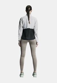Hellgraue und schwarze Sportjacke mit taillierter Passform, kombiniert mit beigen Leggings und hellen Sneakers, die eine schlanke Silhouette zeigen.