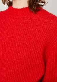 Raudonas mezginys džemperis su grubi tekstūra, aukštu kaklu ir švelniu apdaila. Detalė rodo audinio pynimą ir šiltą spalvą.