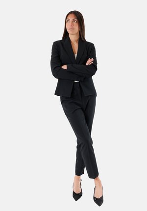 Blazer nero su misura e pantaloni coordinati, abbinati a tacchi a punta neri. Linee pulite, vestibilità strutturata, tessuto liscio e dettagli minimi.