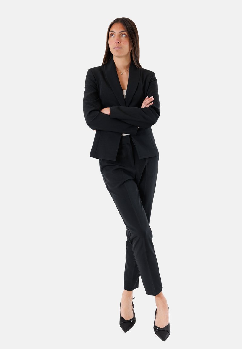 Blazer nero su misura e pantaloni coordinati, abbinati a tacchi a punta neri. Linee pulite, vestibilità strutturata, tessuto liscio e dettagli minimi.