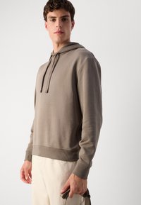 Hettegenser i lys taupe farge, laget av myk stoff med ribbede mansjetter og nederkant. Har snor og en avslappet passform.