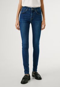 Jeans de denim azul de corte ajustado con un lavado oscuro, cintura de media altura y cinco bolsillos. Presenta costuras en contraste y una ligera textura elástica.