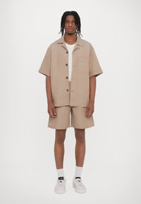 Kortärmad beige linne skjorta med bröstficka, kombinerad med matchande shorts. Modellen bär vit t-shirt, strumpor och ljusa sneakers.