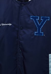 Mörkblå varsityjacka med en framträdande broderad blå "Y"-logotyp och vita detaljer, med tryckknappar framtill.