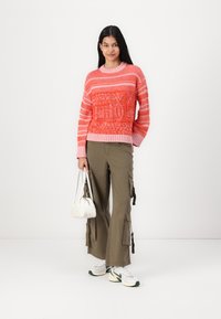 Knitted sweater in pink and orange with patterned textures, paired with olive cargo pants featuring side pockets and a white handbag.