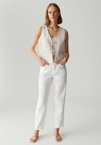 Lichtbeige mouwloze vest met een strikdetail aan de voorkant en twee zakken, gecombineerd met witte rechte broek en minimalistische sandalen.