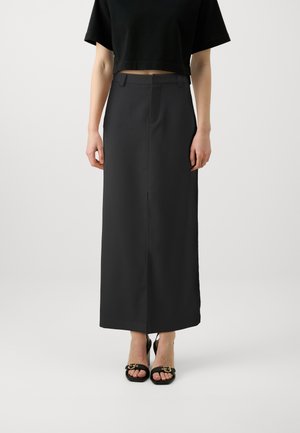 Femme portant une jupe noire taille haute, longueur cheville, avec une fente à l'avant, un top court noir, et des sandales à talons noires avec boucles dorées.