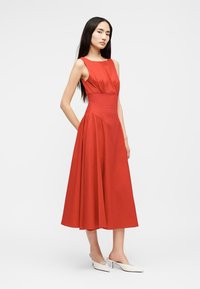 Γυναίκα με μακριά σκούρα μαλλιά, φορώντας κόκκινο μίντι φόρεμα χωρίς μανίκια και λευκά μυτερά τακούνια, στέκεται μπροστά σε ένα απλό ανοιχτόχρωμο φόντο.