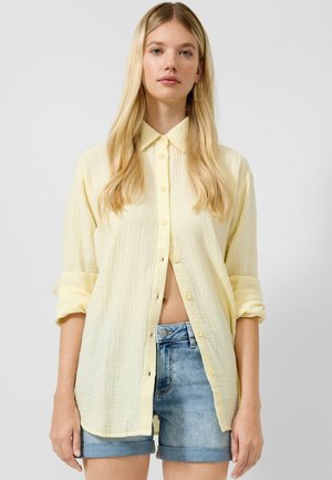 Junge Frau mit langen blonden Haaren, die ein hellgelbes Hemd mit Knöpfen und aufgekrempelten Ärmeln sowie blaue Jeansshorts vor einem einfarbigen Hintergrund trägt.