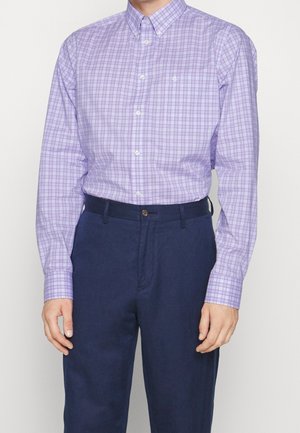 Homme portant une chemise à carreaux lavande à manches longues rentrée dans un pantalon bleu foncé, debout devant un fond uni.