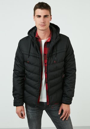 Black puffer jacket with hood, featuring a zippered front and elastic cuffs, worn over a red plaid shirt and white t-shirt.