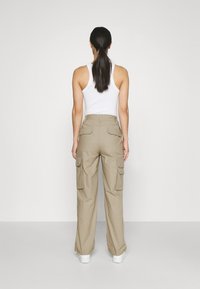 Žena s dlouhými tmavými vlasy v culíku, oblečená v bílé tílko, béžových cargo kalhotách s několika kapsami a bílých teniskách, stojící proti jednoduché zdi.