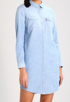 Kobieta ubrana w jasnoniebieską, długorękawową sukienkę koszulową z denimu z kieszeniami na piersi i kołnierzykiem, stojąca na jednolitym tle.
