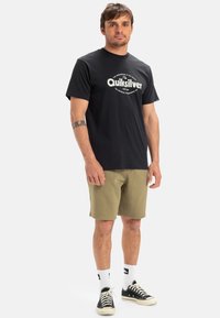 Černé bavlněné tričko s logem Quiksilver, krátké rukávy, v kombinaci s olivově zelenými šortkami, bílými ponožkami a černými teniskami. Jednoduchý design.