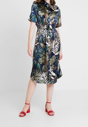 Femme portant une robe bleu marine imprimée feuilles tropicales, boutonnée devant, avec une ceinture à la taille et des manches courtes, associée à des sandales rouges à talons blocs.