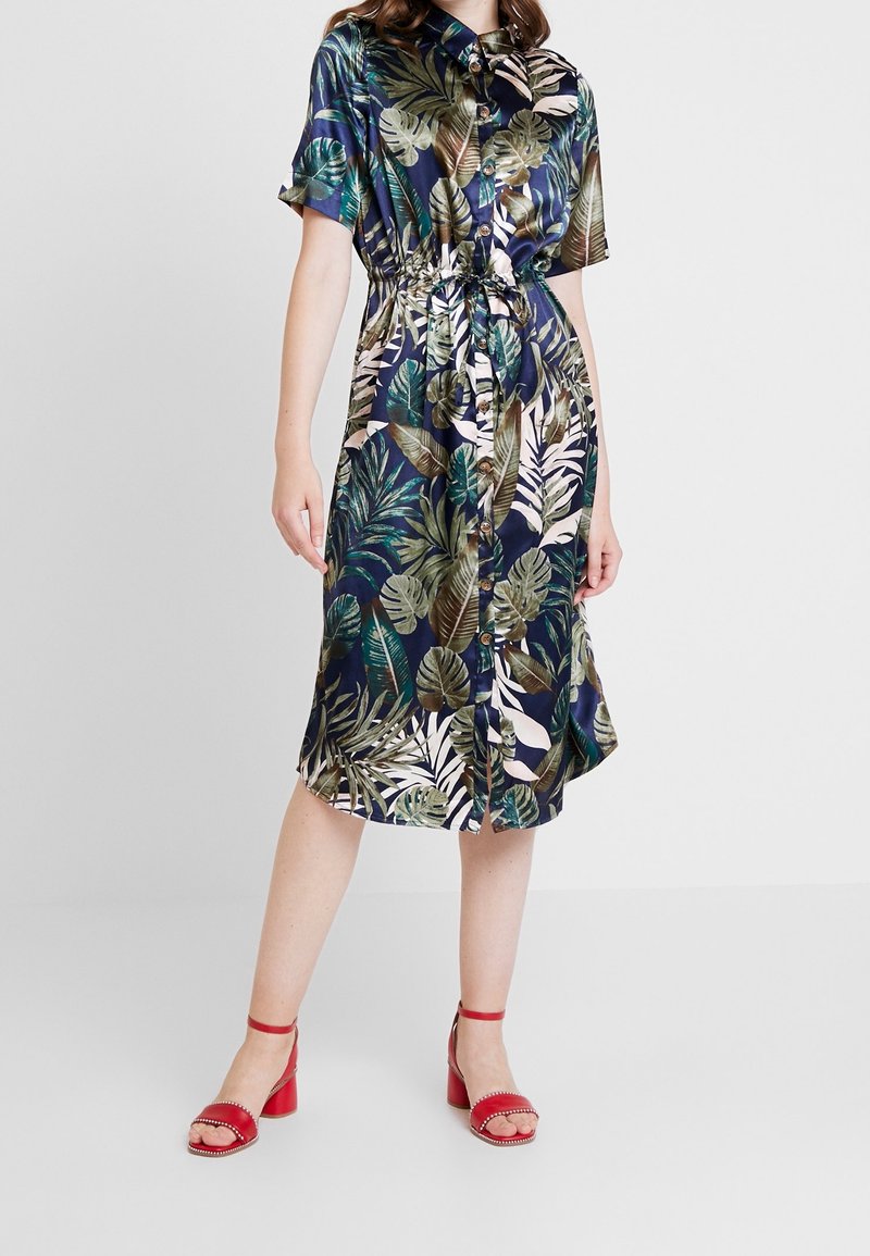 Femme portant une robe bleu marine imprimée feuilles tropicales, boutonnée devant, avec une ceinture à la taille et des manches courtes, associée à des sandales rouges à talons blocs.