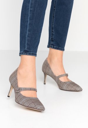 Fødder iført spidse Mary Jane højhælede sko med ternet mønster kombineret med mørkeblå slim fit jeans mod en ensfarvet hvid gulv og væg.