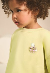 Ljusgul sweatshirt med ett runt, mönstrat äppelformad patch som har leende ögon, grön blad och rosa detaljer.