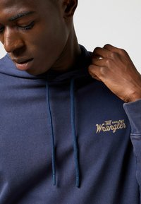 Felpa blu navy con tessuto a trama, caratterizzata da un logo stampato in oro "The Wrangler" e dettagli con cordini. Dettaglio della parte superiore del corpo.
