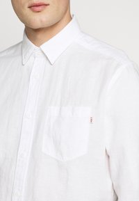 Homme portant une chemise blanche à boutons avec un col pointu et une poche poitrine étiquetée "HKT" en lettres rouges.