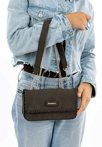 Brun faux læder håndtaske med kædetras, prydet med flettet trim og en metal logo-plade, heldt af en person iført en denimjakke.