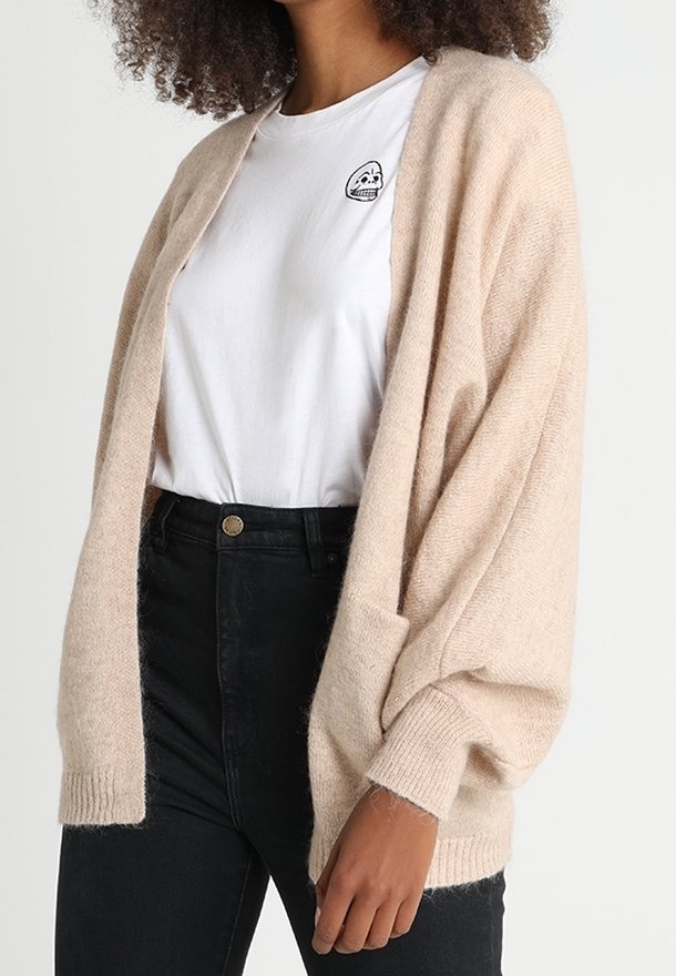 Beige, oversize svetr vyrobený z měkkého materiálu s širokými rukávy a žebrovanými manžetami, nošený přes bílé tričko a černé džínsy s vysokým pasem.