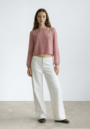 Giovane donna in piedi con indosso un top corto rosa a maniche lunghe con una spalla scoperta, pantaloni bianchi a gamba larga e scarpe marroni su uno sfondo semplice.