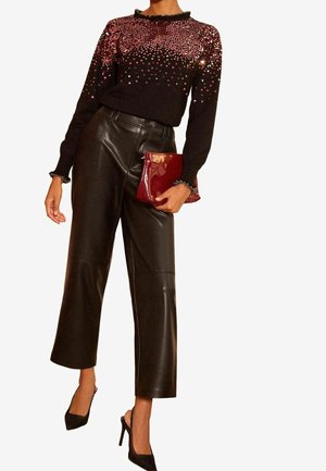 Femme portant un haut noir à manches longues avec des paillettes roses, un pantalon court en cuir noir, des talons hauts noirs, tenant une pochette rouge vernie.