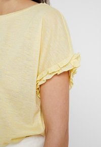 Žlutý krátký rukáv top s jemnou texturou a volánkovými okraji. Materiál vypadá lehký a mírně průsvitný, s ležérním designem.