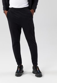Zwarte sportbroek met een taps toelopende fit, gemaakt van rekbare stof, met een elastische tailleband en zijzakken met rits, gecombineerd met zwarte sneakers.