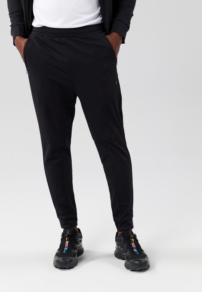 Zwarte sportbroek met een taps toelopende fit, gemaakt van rekbare stof, met een elastische tailleband en zijzakken met rits, gecombineerd met zwarte sneakers.