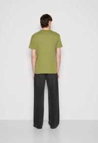 Camiseta de algodón verde oliva con mangas cortas, diseño ajustado, vista desde la parte de atrás, combinada con jeans negros de pierna ancha.