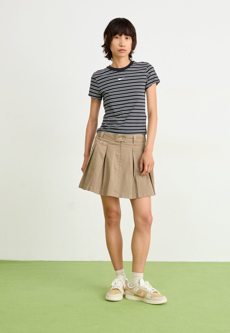 Pasiasta biało-czarny t-shirt z krótkim rękawem zestawiony z beżową plisowaną spódnicą. Modelka nosi beżowo-białe sneakersy z jasnymi skarpetkami. Tło w kolorze zielonym.
