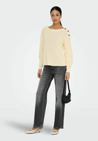 Femme debout portant un pull en tricot crème avec des boutons, un jean noir délavé, des talons beiges, des boucles d'oreilles créoles et tenant un petit sac à main noir.