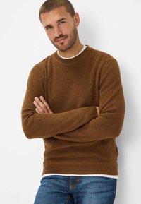 Bruine geribde trui met een ronde halslijn en lange mouwen, gedragen over een wit shirt, in combinatie met een spijkerbroek.
