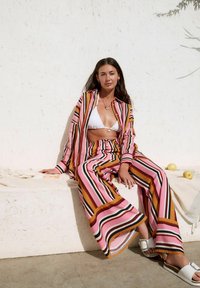 Pantaloni a gamba larga a righe rosa, arancioni e neri con camicia lunga abbinata, indossati con un top bikini bianco e sandali bianchi.