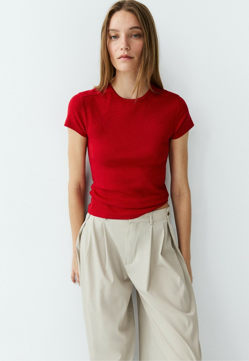 Woman with long straight hair wearing a fitted red short-sleeve top and loose beige pleated trousers against a plain white background.