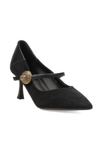 Zapato de tacón alto de ante negro con punta afilada, correa ajustable y adorno metálico redondo. Cuenta con un tacón con forma de cono esculpido.