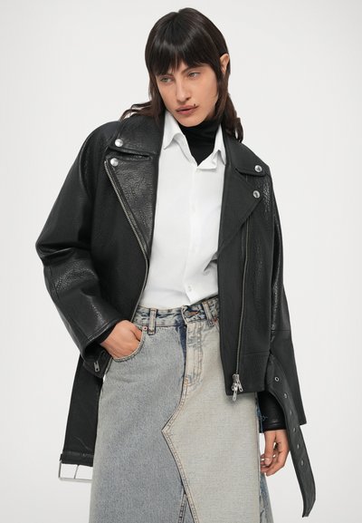 Casaco de couro preto para motociclista com acabamento texturizado, ferragens prateadas e design assimétrico, usado por cima de uma camisa branca e uma saia de ganga.