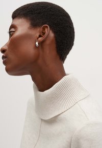 Neutralkleurige trui met een hoge ribbelkraag. Model draagt een stijlvolle, glanzende zilveren oorring. Achtergrond is eenvoudig en licht.