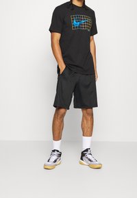 Camiseta negra de manga corta de Nike con un logo de cuadrícula azul y amarillo, combinada con pantalones cortos negros y calcetas blancas, acabada con zapatillas deportivas negras y grises.