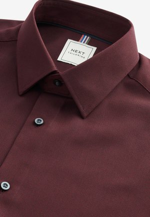 Burgunderfarbene Button-Down-Bluse aus strukturiertem Stoff, mit einem strukturierten Kragen, zwei schwarzen Knöpfen und einem Etikett mit bunten Streifen.