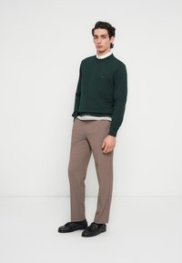 Πράσινο φούτερ με στρογγυλή λαιμόκοψη φορεμένο πάνω από ένα λευκό πουκάμισο με μαύρες ρίγες, σετ με καφέ παντελόνι ραμμένο και μαύρα παπούτσια.