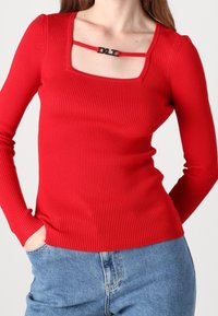 Rotes, geripptes Langarmoberteil mit quadratischem Ausschnitt und einem schwarzen Akzentband. Trägt dazu eine hochgeschnittene blaue Jeans.