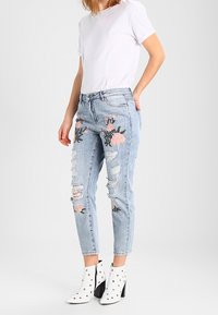 Světle modré oprané džíny s květinovou výšivkou v růžové a zelené, nošeny s bílým tričkem a bílými kotníkovými botami se cvočky.