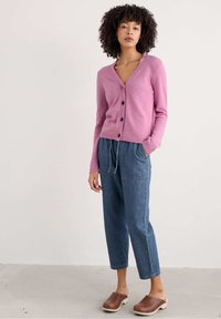 Roze gebreide cardigan met V-hals, zwarte knopen en geribbelde zoom, gecombineerd met lichtblauwe baggy spijkerbroek en bruine klompen.