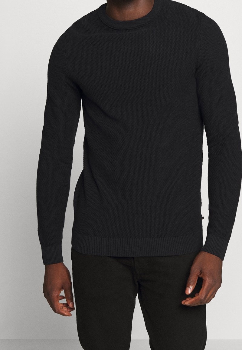Pull noir en tricot à col rond avec tissu texturé, manches longues, et poignets et ourlet côtelés. Associé à un pantalon foncé à coupe décontractée.