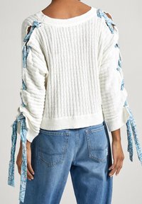 Maglione bianco lavorato a maglia con texture a coste, spalle scoperte fissate con nastri floreali blu, abbinato a jeans in denim blu a vita alta.