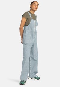 Salopette en denim rayée avec fermeture éclair à l'avant, deux poches avant et jambes larges, associée à un t-shirt vert ajusté et des chaussures de sport.