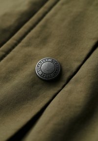 Tissu vert avec une finition texturée présentant un bouton en métal rond marqué "Superdry Merchant Store" en lettres noires.